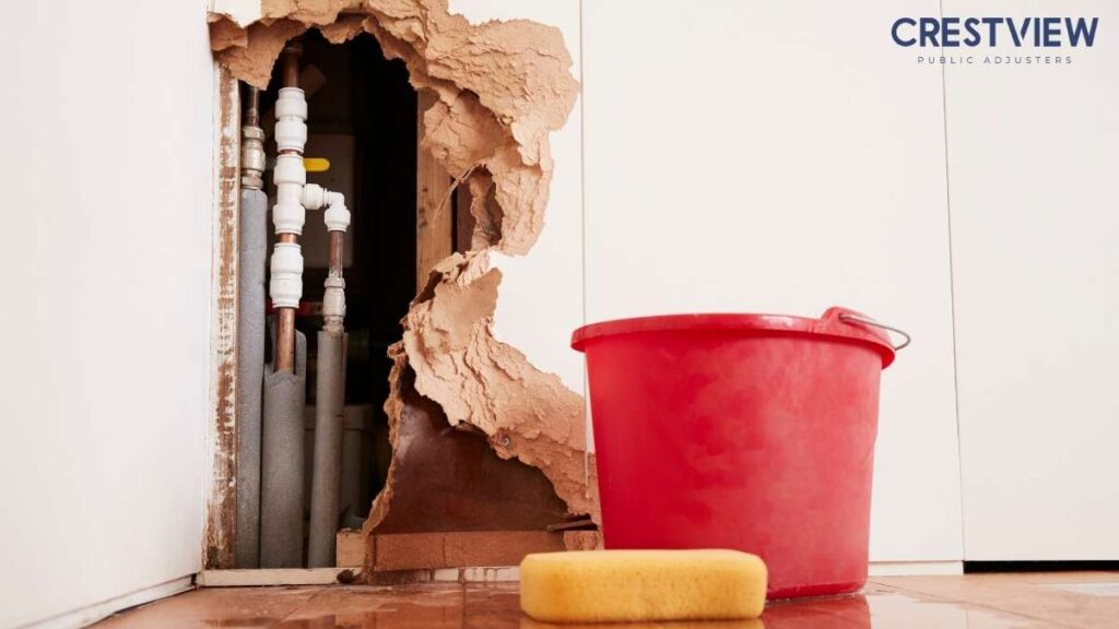 Commercial Water Damage Claim In New Jersey: Why Business Owners Should Work With A Public Adjuster 1 Burst pipe behind torn drywall exposing interior plumbing, with a red bucket on a wet floor